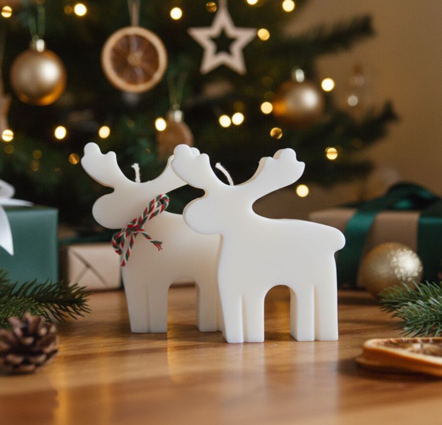 Świece sojowe w kształcie jelonków stojące na drewnianym stole, z choinką i świątecznymi prezentami w tle. Białe dekoracyjne świece Candelovo, zimowy klimat, naturalne bożonarodzeniowe ozdoby.